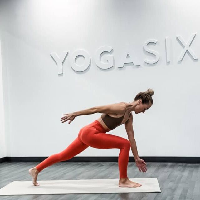 Woman demonstrates yoga pose in yoga studio with words Yoga Six embossed on wall behind her