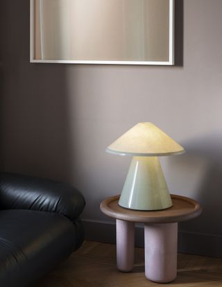 A glass lamp by Umberto Riva consisting on two stacked cones in light green. The lamp is shown on a pink side table next to a leather upholstered sofa