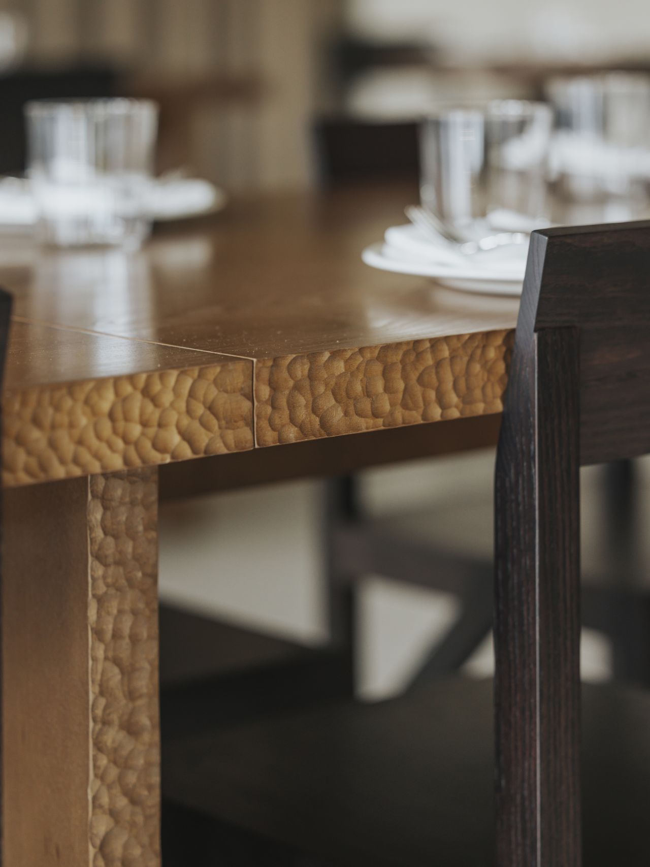 A restaurant with brutalist interiors features wooden upholstered banquettes with warm, brown fabric matching with matte chrome tables with a geometric shape.