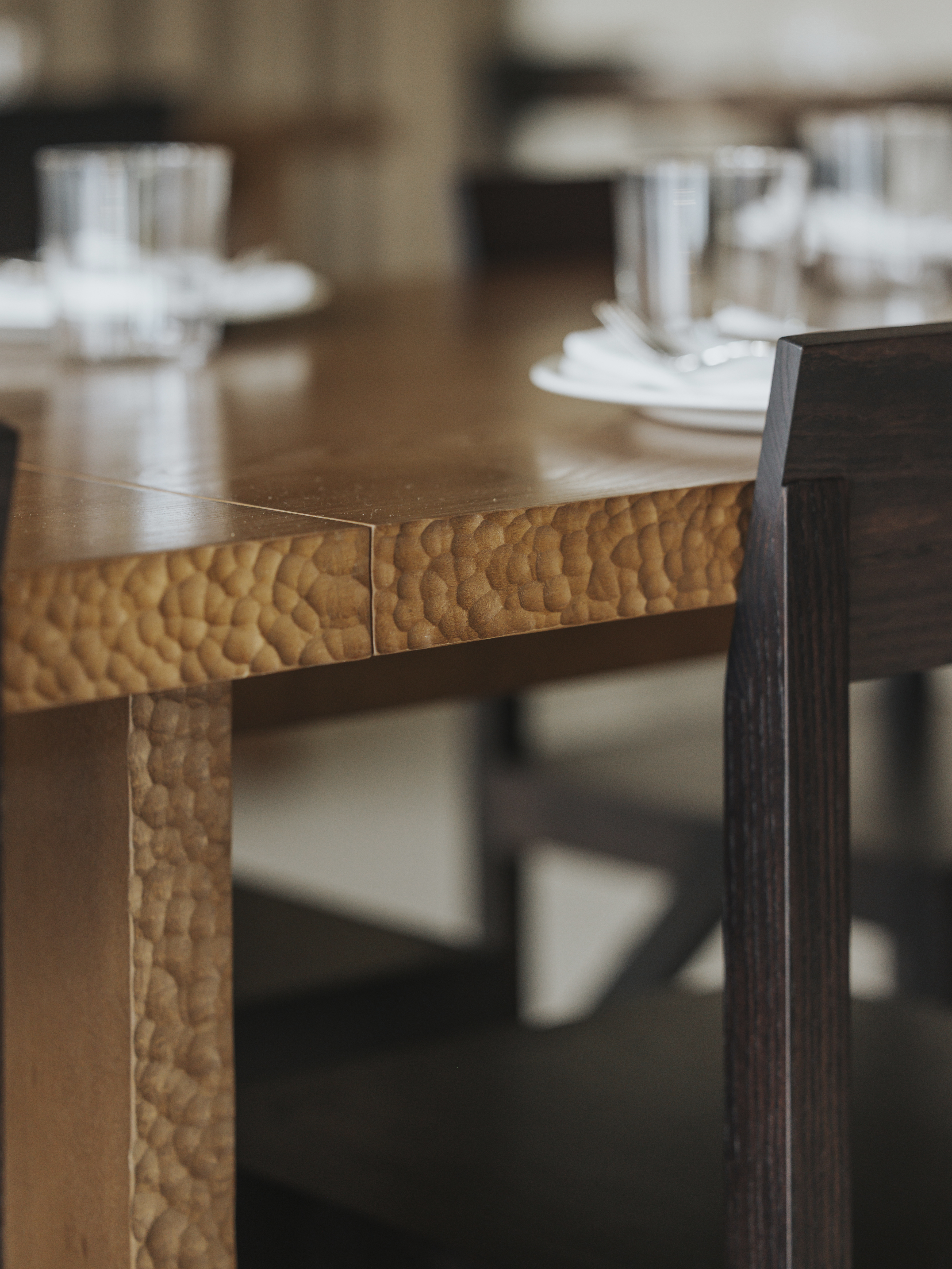 A restaurant with brutalist interiors features wooden upholstered banquettes with warm, brown fabric matching with matte chrome tables with a geometric shape.