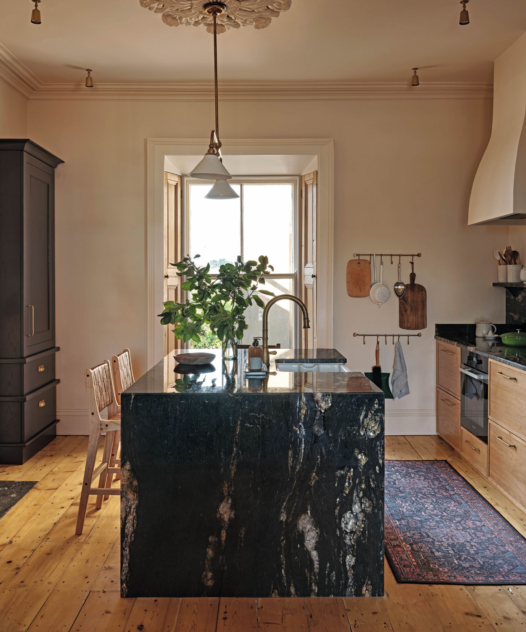 a warm timber kitchen with a dark stone island built into a victorian home with plaster ceiling rose, cooker hood and small pendant lights