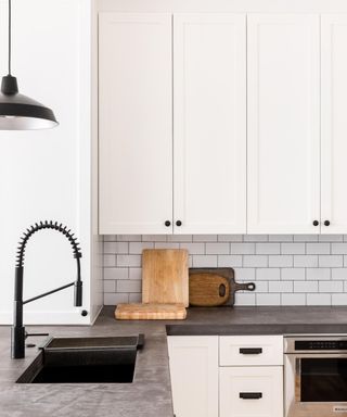 A kitchen with white cabinets, white subway splashback with four chopping boards leaning against it, dark gray countertops, a black sink and faucet to the left, and white cabinets with drawers and an oven below