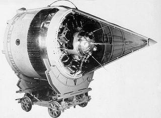 A conical spacecraft on four wheels on its side, with a cone fairing cut in half to reveal a smaller chrome antenaed space probe.