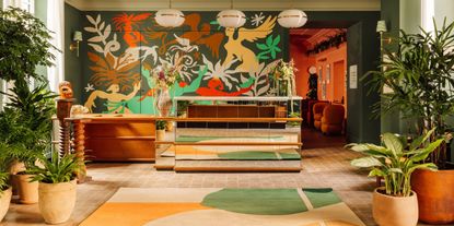 A colorful hotel lobby, filled with trailing plants in terracotta pots and decorated with handmade colorful murals and matching rugs, boasts a wood and mirror reception with sculptural lighting pending above it.