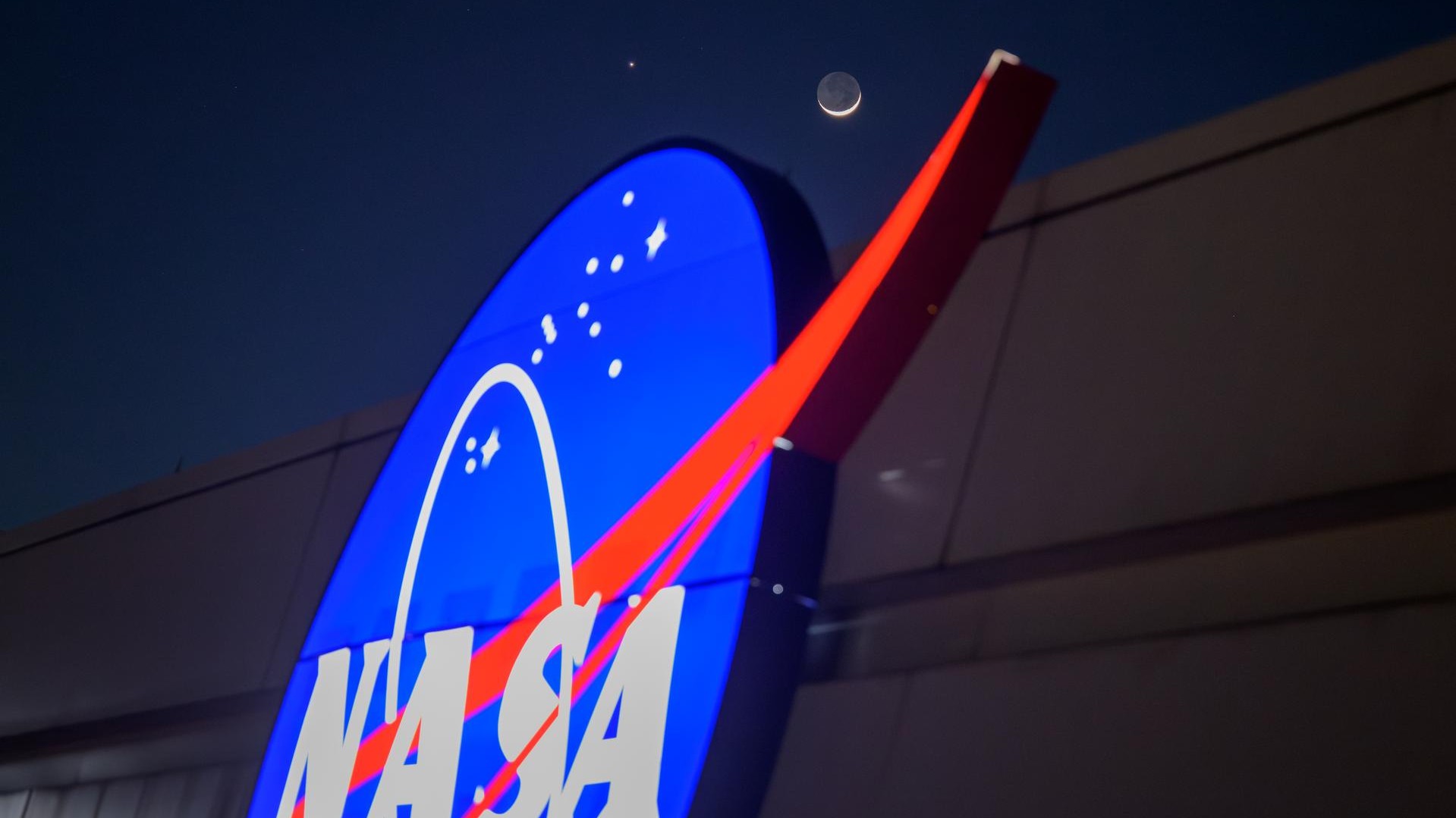 The crescent moon and Venus shine in the night sky above the NASA "Meatball" logo on the side of a building.