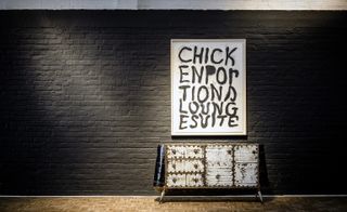 View of framed text art on the wall and a distressed white and brown cabinet beneath it in a space with black painted brick walls and brown patterned flooring
