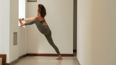 woman wearing khaki leggings and crop top doing a standing leg stretch up a wall in a white wall space 