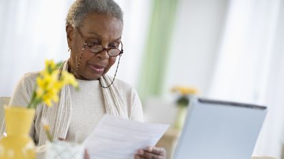 An older woman looks over a bill while sitting at her kitchen table.