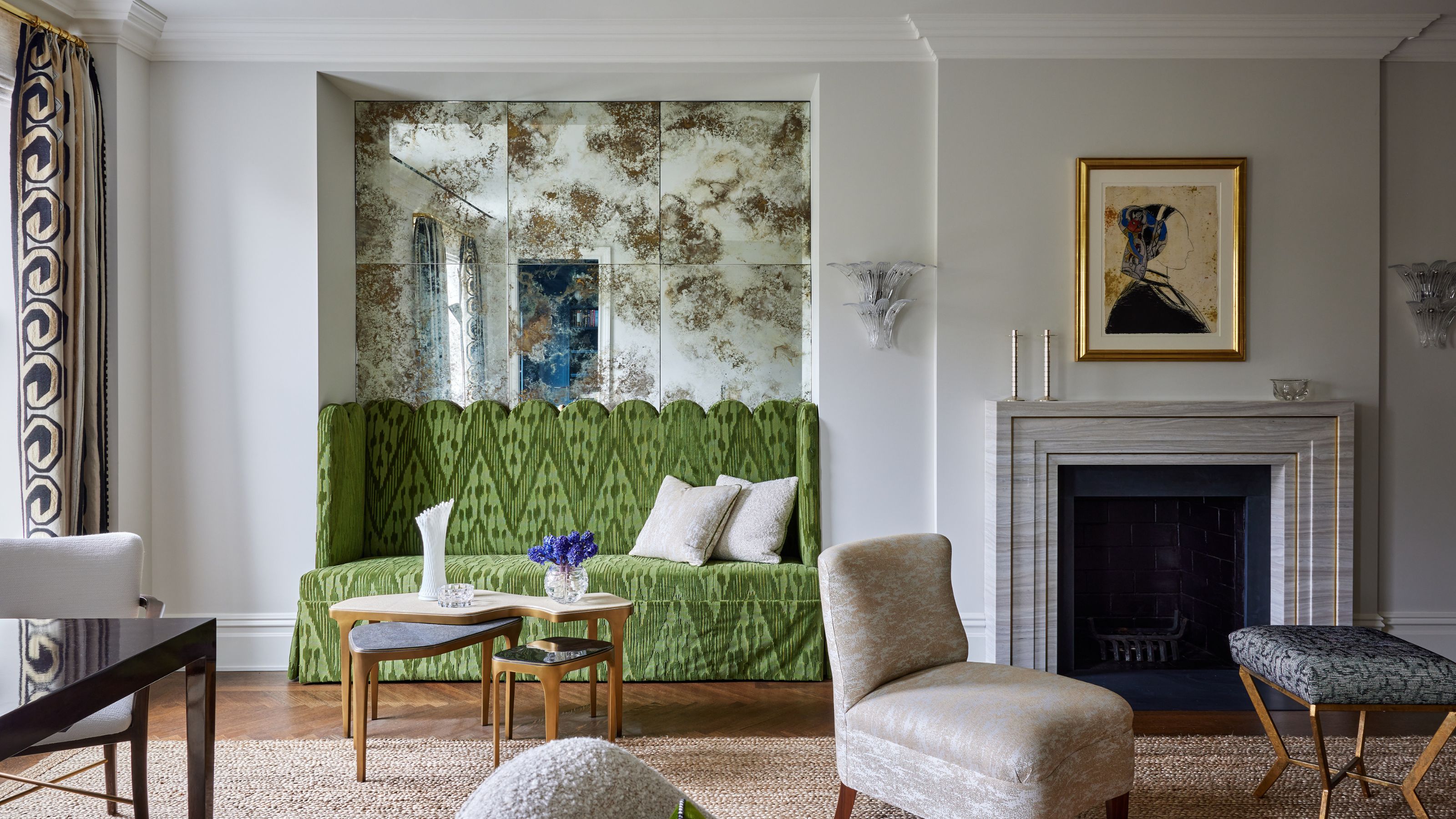 living room with marble fireplace with artwork hung above, a stool and armchair, a banquette seat upholstered in green with a mirrored wall behind it, patterned curtains, timber floors, jute rug, and nesting coffee tables in front of banquette seat