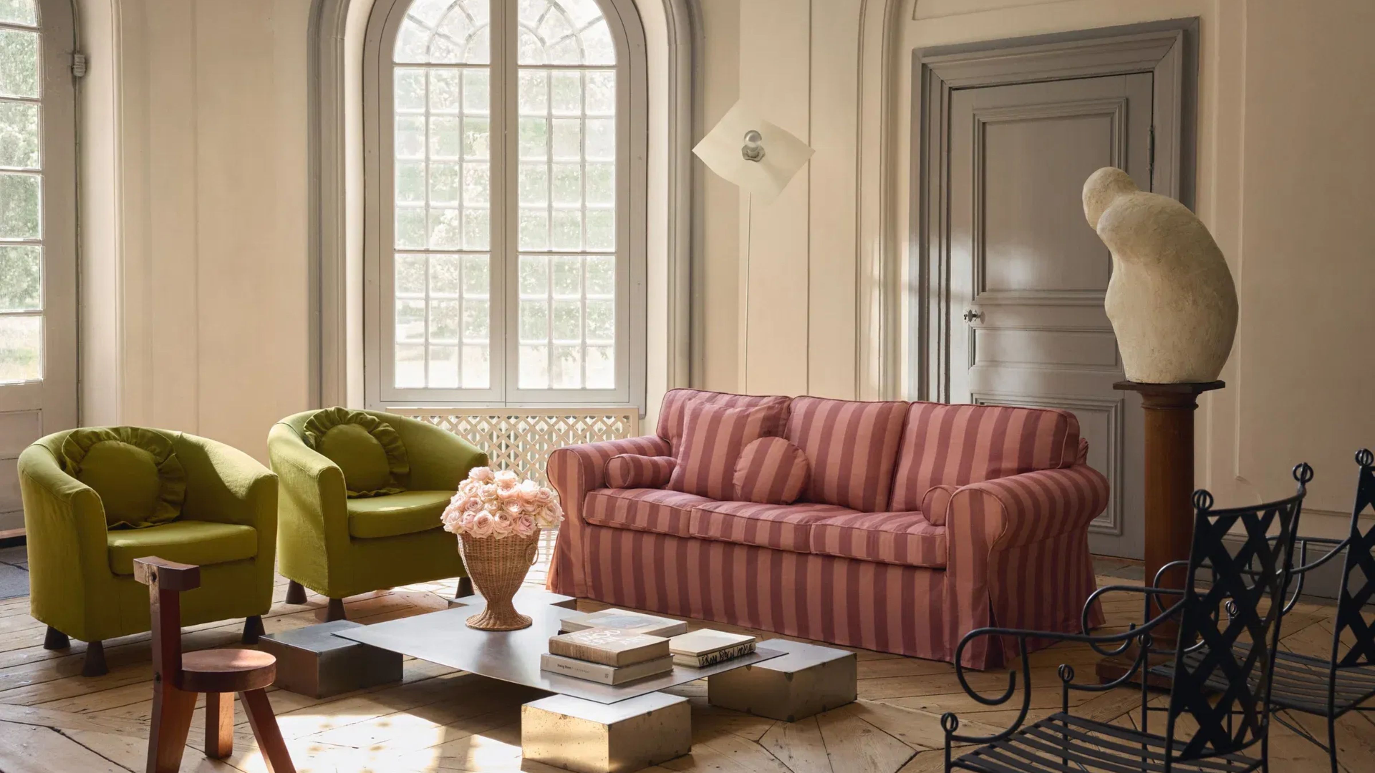 Sunlit living room with wooden floors and arched windows featuring two green accent chair alongside a tonal pink-striped, slipcovered IKEA sofa