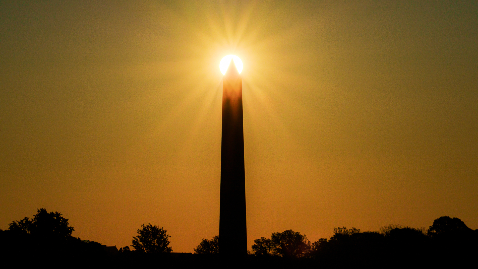 The sun is photographed shining behind the Washington Monument at sunset.