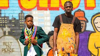 LICCLE BIT (SAMSON) in school uniform and MANJARO (BUNMI OSADOLOR) in paint splattered overalls standing in front of a wall painted with cartoons