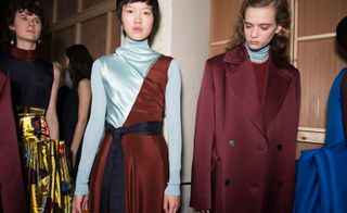 Model wears a high-neck silk dress in blue and maroon, whilst another wears a tailored dark-red trench coat