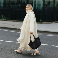 Woman wears fringed scarf coat, black bag. 