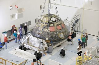 technicians surround a silvery space capsule inside a high-ceilinged, white-walled room.