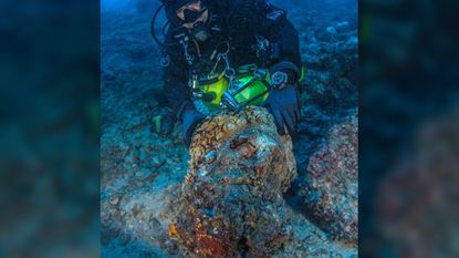 This massive marble head found by the latest excavations at the Antikythera wreck site is thought to represent the Greek demigod Heracles. Here we see a diver underwater examining a very worn marble sculpture head.