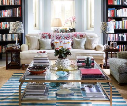 Living room with bay window, pale blue walls, built in bookcases, cream sofa with multi-coloured cushions and large square coffee table