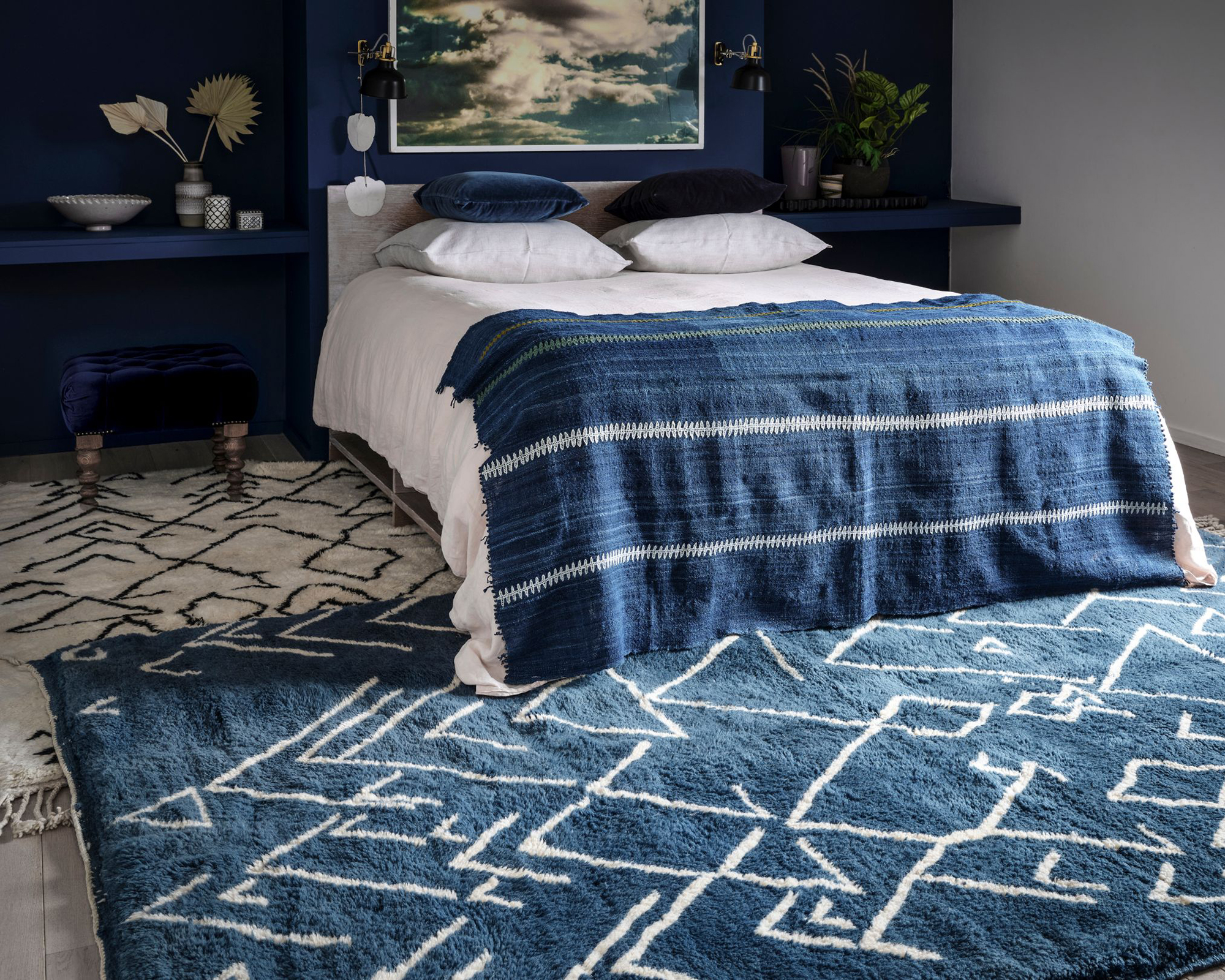 A blue bedroom with a double bed, floating shelves and layered Berber-style rugs