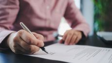 A person signs a document at a desk, only their hands and torso showing.