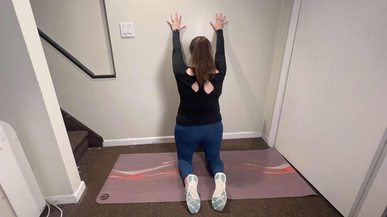 Trainer Jennifer RIzzuto performs a back mobility stretch on the floor. She is kneeling close to a wall on a yoga mat, with both hands on the wall in front of her.
