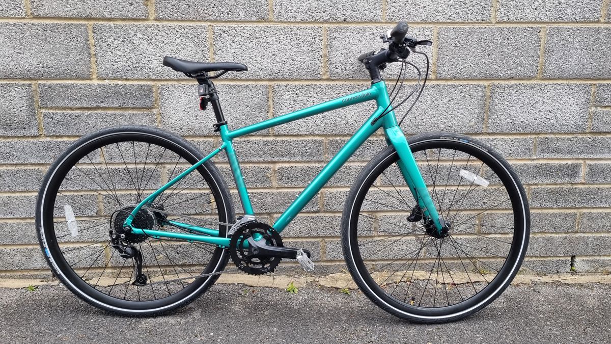 A metallic green flat bar Cannondale Quick 3 bike leaning against a grey brick wall