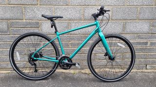 A metallic green flat bar Cannondale Quick 3 bike leaning against a grey brick wall