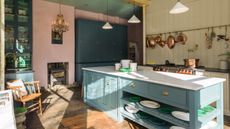 kitchen with pink wall with stove, teal cabinetry, cream shiplap wall with brass hanging rail and copper saucepans, large kitchen island painted in teal, white countertop, wooden floorboards, alcove storage with glazed doors, wooden chair, chandelier and enamel pendant lights