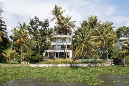 The Plumeria a concrete indian house designed as stacked boxes peaking behind palm trees and tropical planting across a lake