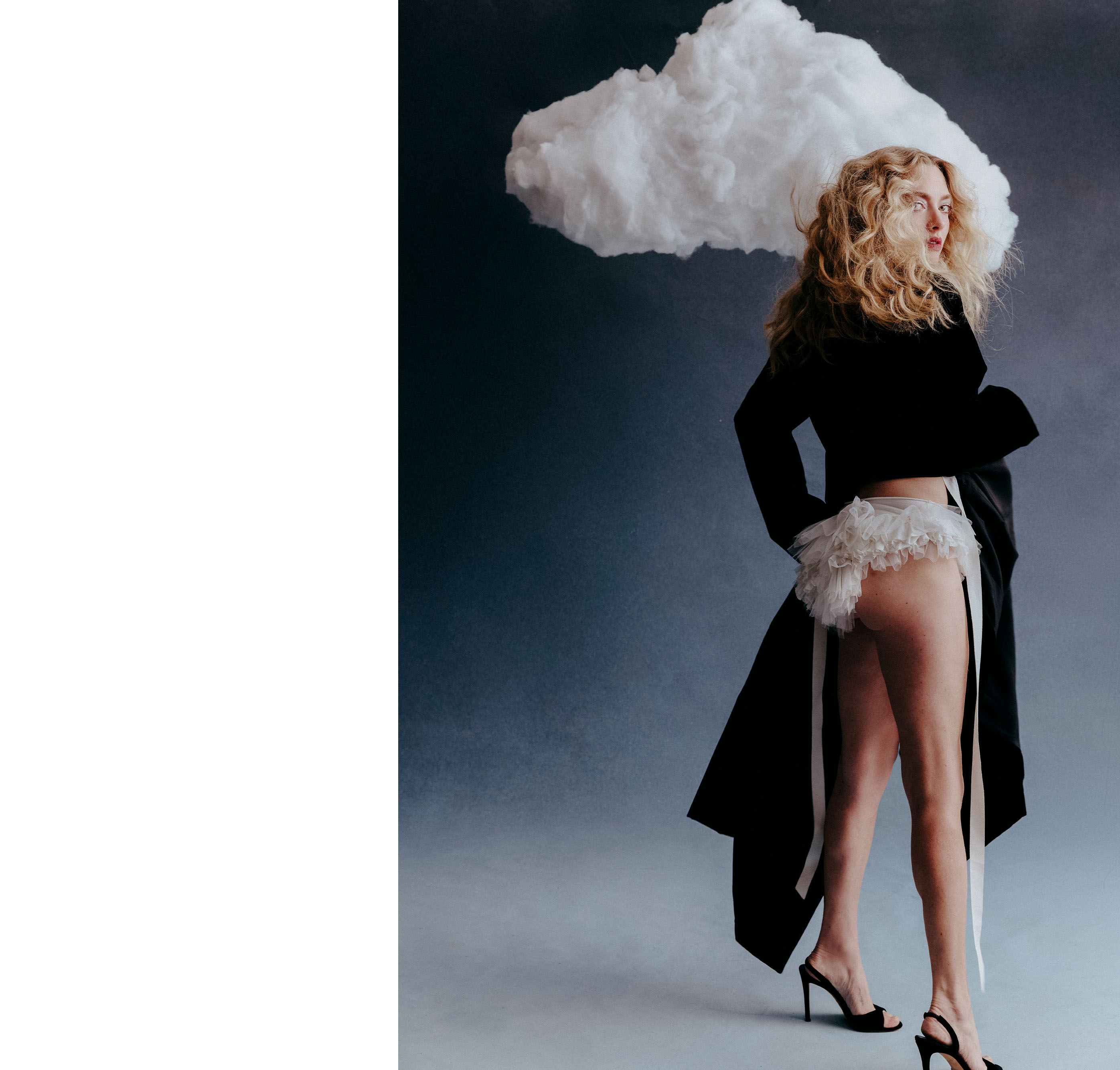 Amanda Seyfried for Who What Wear&#039;s December cover story. She is photographed standing in front of a hanging cloud. She is looking back at the camera exposing a pair of white frilly briefs underneath her black coat.