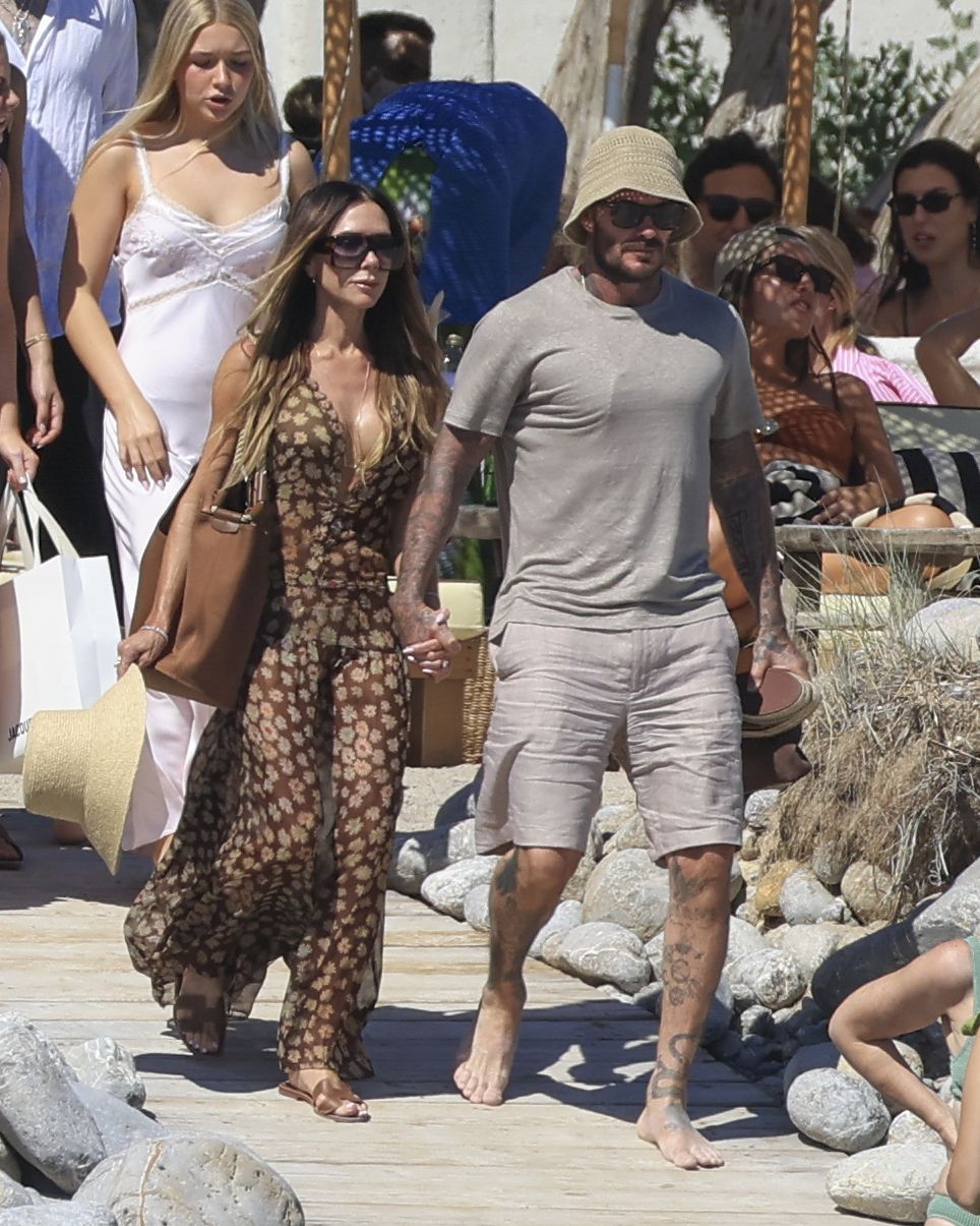 Victoria and David Beckham walk along a beach holding hands. Victoria wears a brown sheer dress with Hermes Oran sandals whilst David wears a grey t-shirt with grey linen shorts.