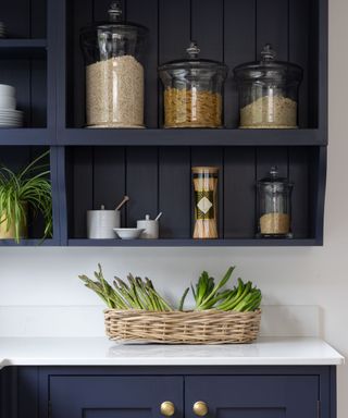 A dark blue open kitchen cabinet with three glass jars with pasta and grains at the top, white ceramic spice jars and two jars at the bottom, and a white countertop with a basket of spring onions on it underneath