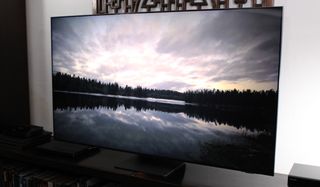 En Samsung QN95A Neo QLED TV står på en svart TV-bänk och visar en bild över en sjö och en skog på skärmen.