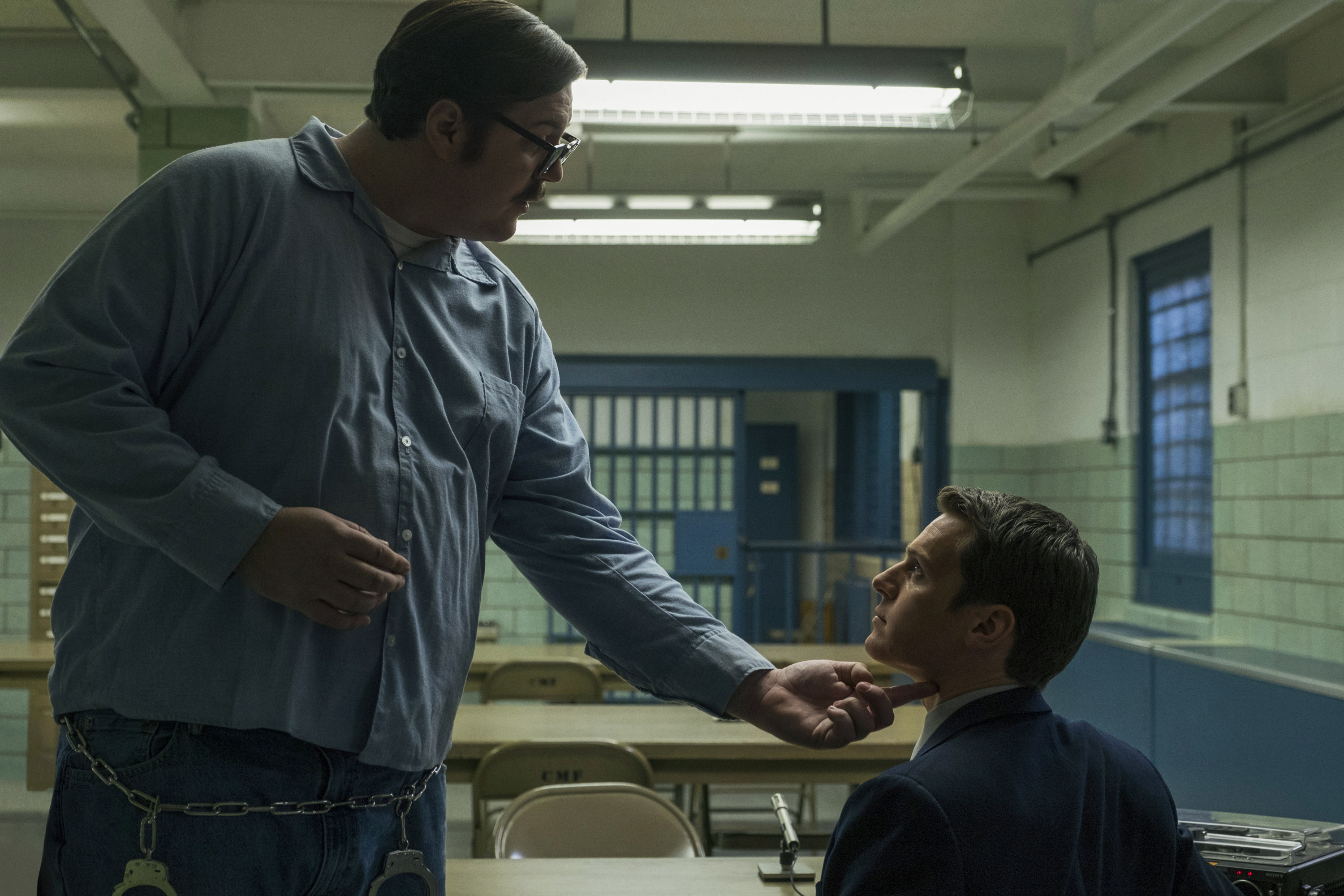 a serial killer in a blue jail jumpsuit reaches down and touches the face of an fbi officer in the show mindhunter