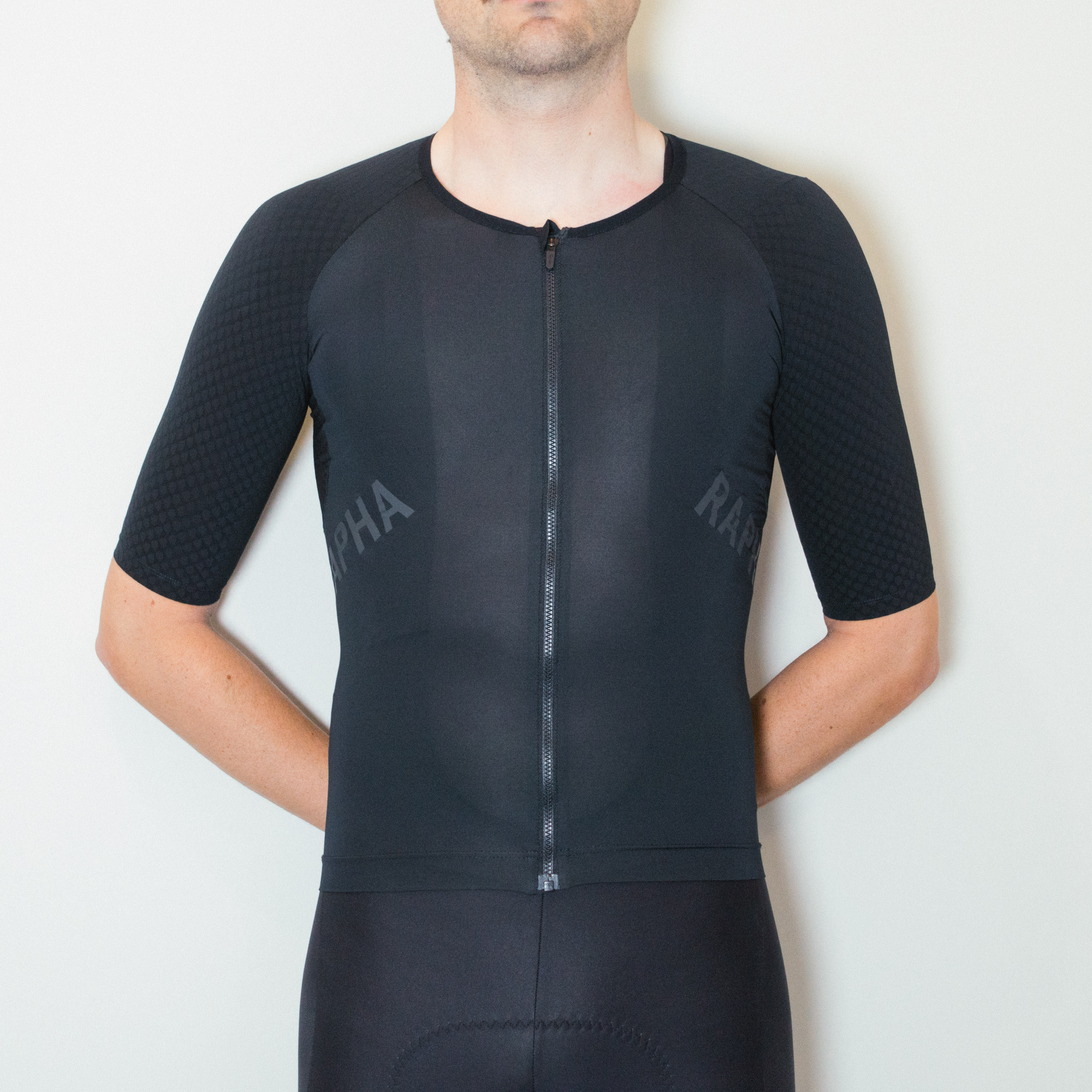 A white man stands against a white wall wearing black cycling shorts and a tight all black jersey