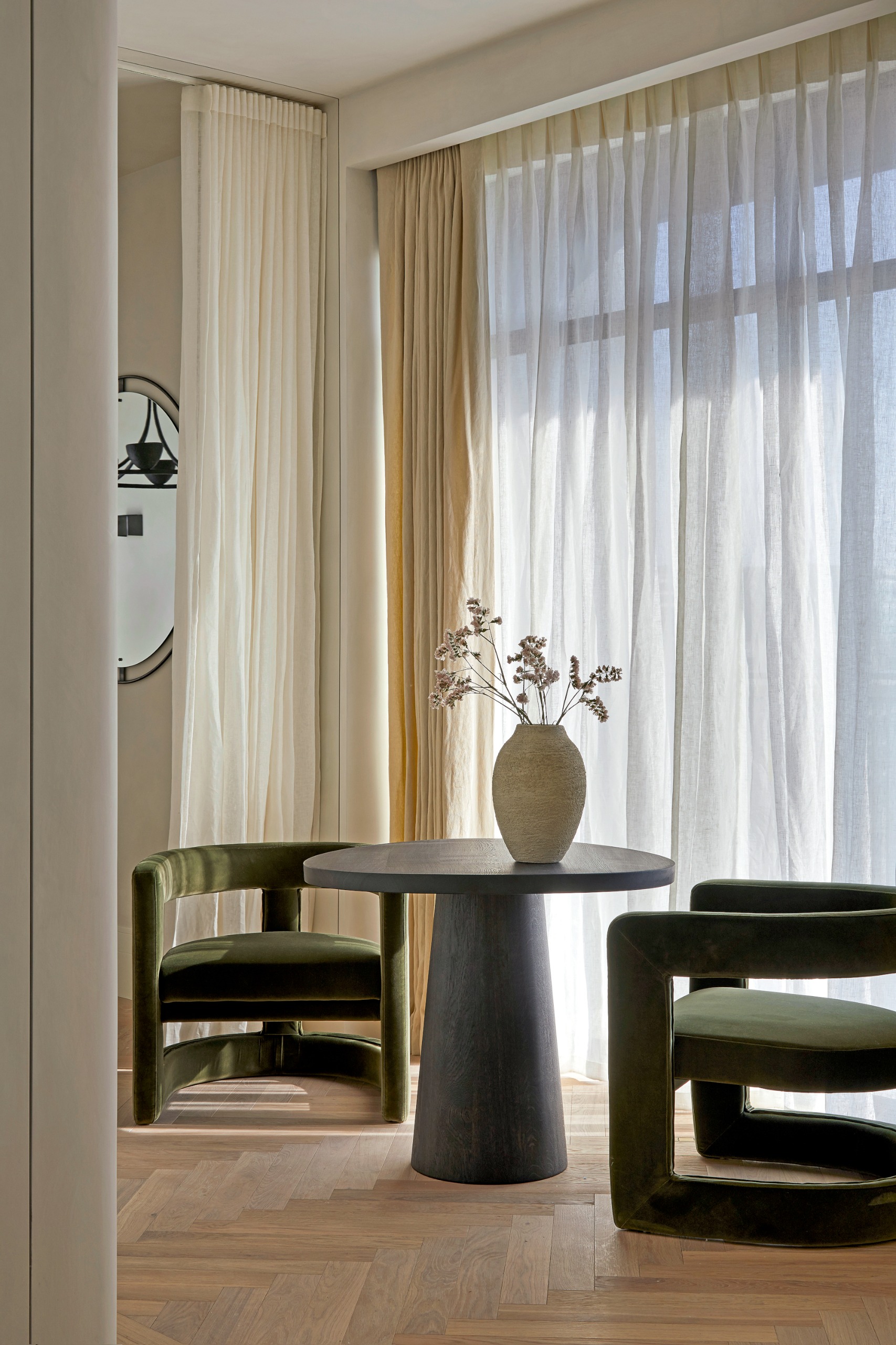 Living room with cream sheer curtains, black round table and olive green velvet armchairs, and parquet oak floor