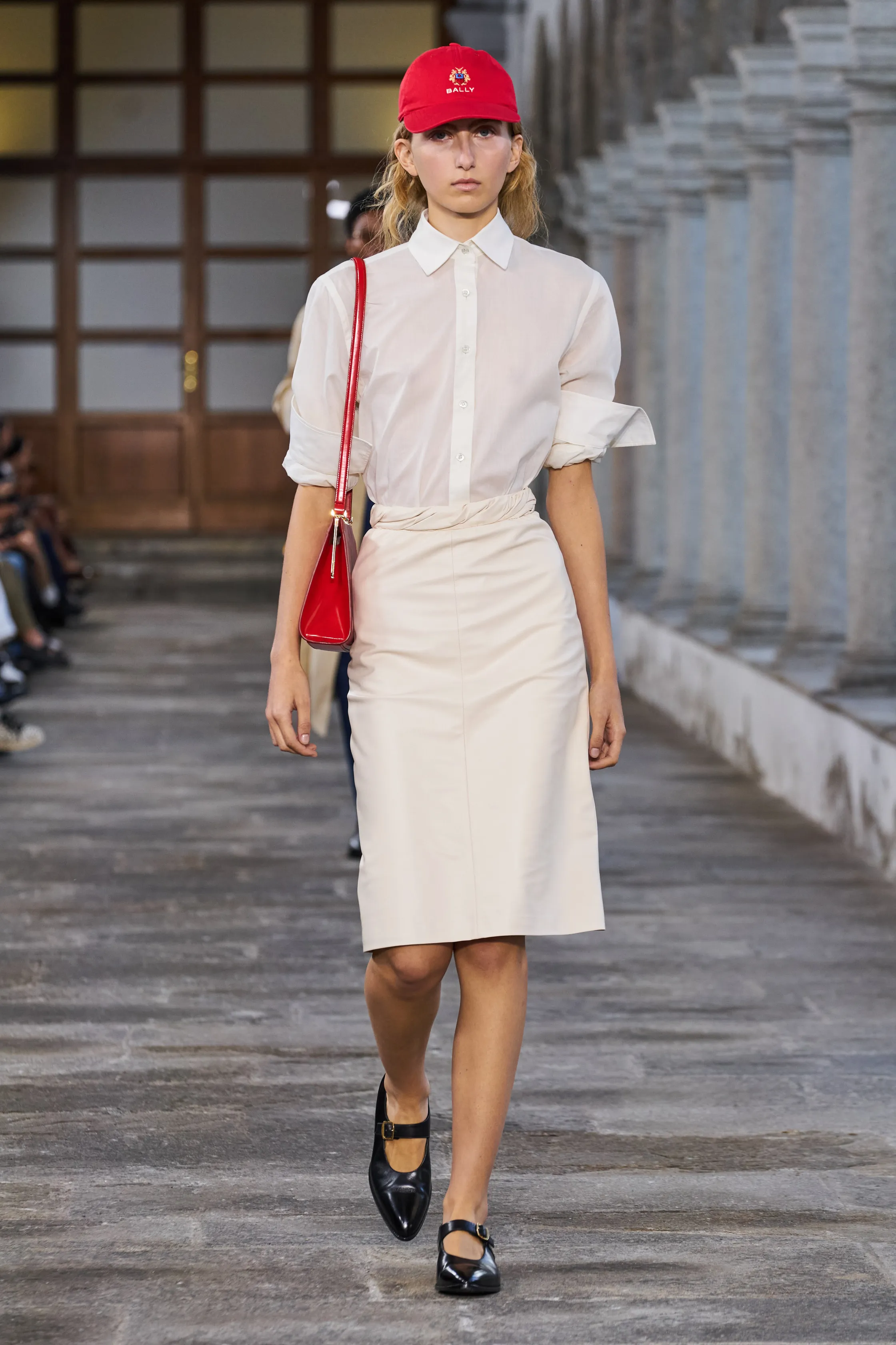 A model wears a white shirt, pencil skirt, red baseball hat, mary jane shoes, and a red bag in Bally Spring 2024.