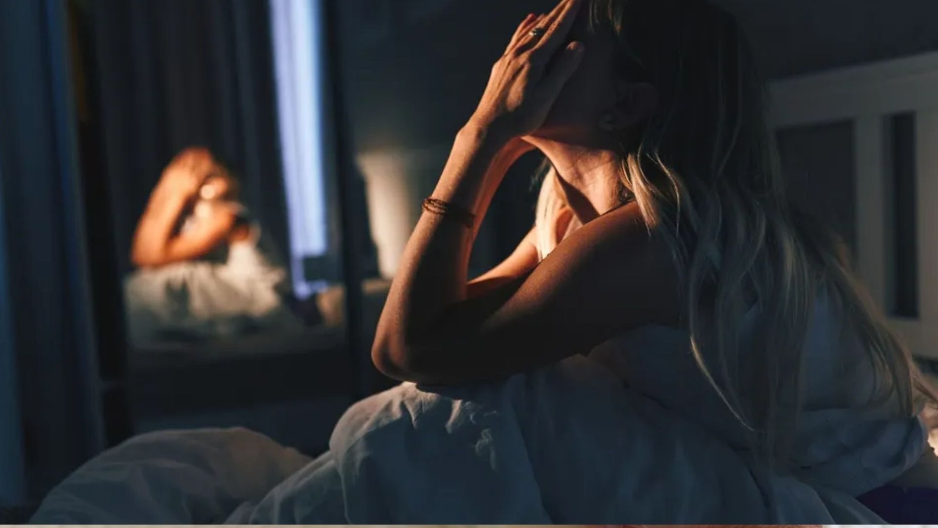 The image shows a blonde haired woman sitting up in bed inside a dark bedroom.  Her knees are bent and her hands are placed over her face. 