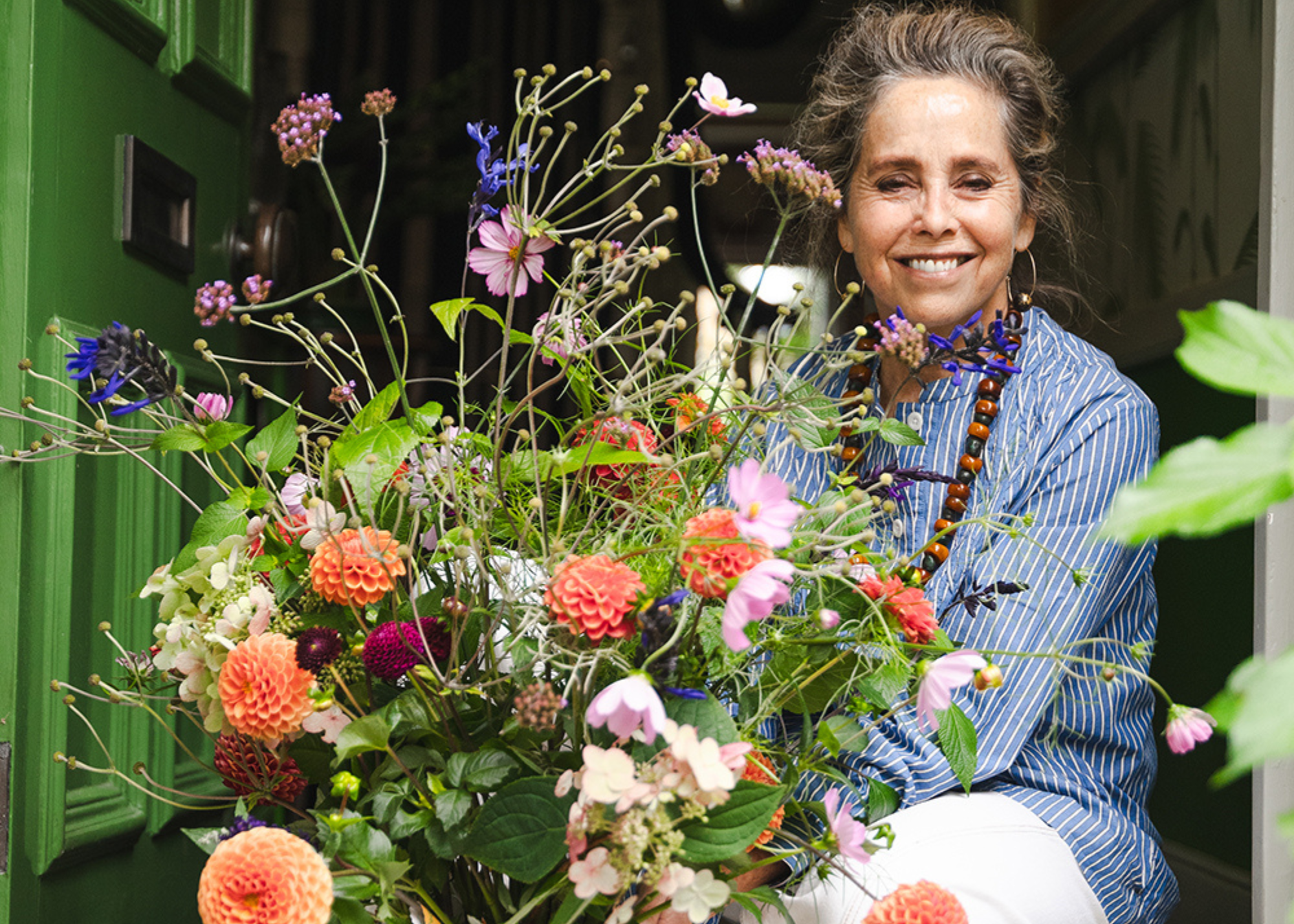 A woman in a striped shirt in a green doorway by a vase of colorful dahlias