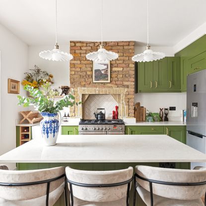 an open-plan kitchen diner with green cabinets and a kitchen island with three cream coloured bar stools and an exposed brick chimney breast with a stainless steel range cooker