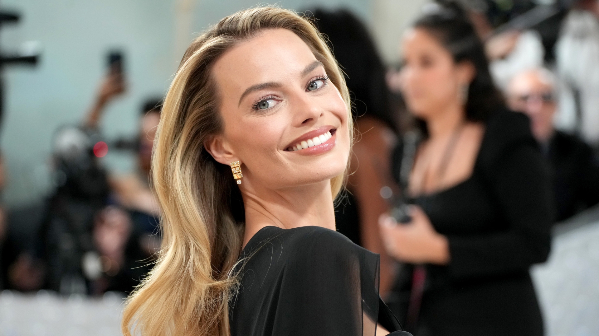 Image of Margot Robbie smiling, looking over her shoulder with long blonde hair and wearing a black gown