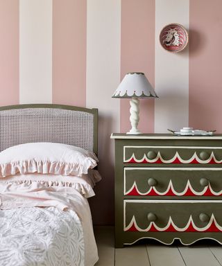 A bedroom with dusky pink and light pink striped wall, a sage green woven bed frame with light pink ruffled bedding, and a dark green chest of drawers with scalloped red decoration and a white table lamp on top of it, plus a red circular plate above this