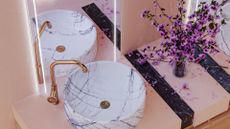 light pink bathroom cabinet with white marble sink and gold tap and light up mirror behind.