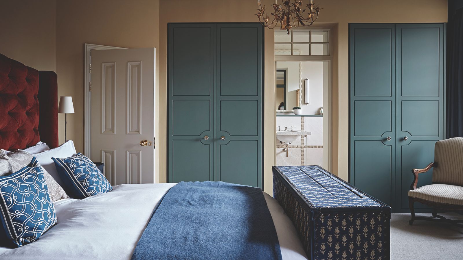 A bedroom with blue built-in closets, a red velvet headboard, white bedding, blue cushions, and a blue storage ottoman at the end of the bed.