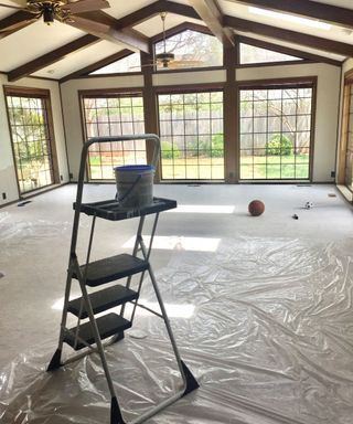Living room with high ceilings and wood beams mid decorating, with a covered over floor and a step ladder
