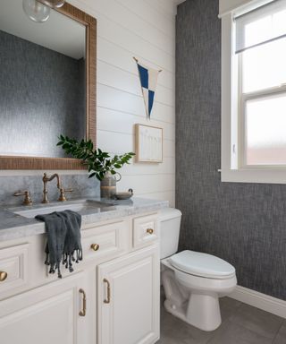 A bathroom with a cream sink unit with gray granite and brushed gold taps, a brown wooden woven mirror, white wall planks with a wall art print and blue and white triangle, a white toilet, and a gray wall with a window