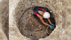 An archaeologist wearing a black and red tank top, white hard hat, and blue gloves excavates a circular pit that contains a skeleton on its side in the fetal position