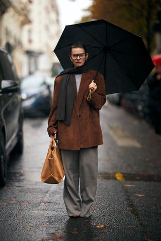 A woman in gray pants, a brown suede blazer, grey top, and brown scarf