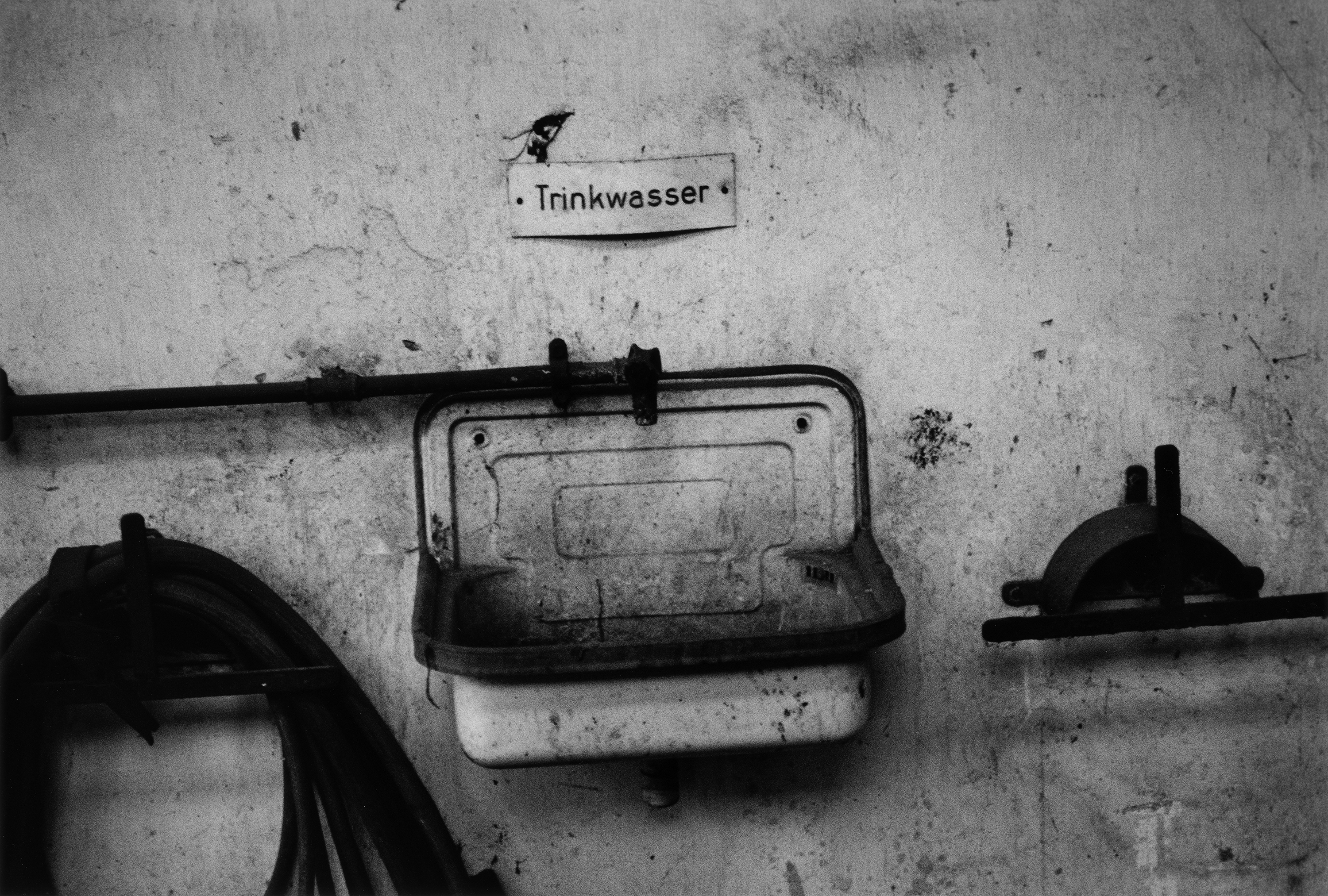 Black and white photograph of drinking tap and basin set in grubby factory wall, by David Lynch