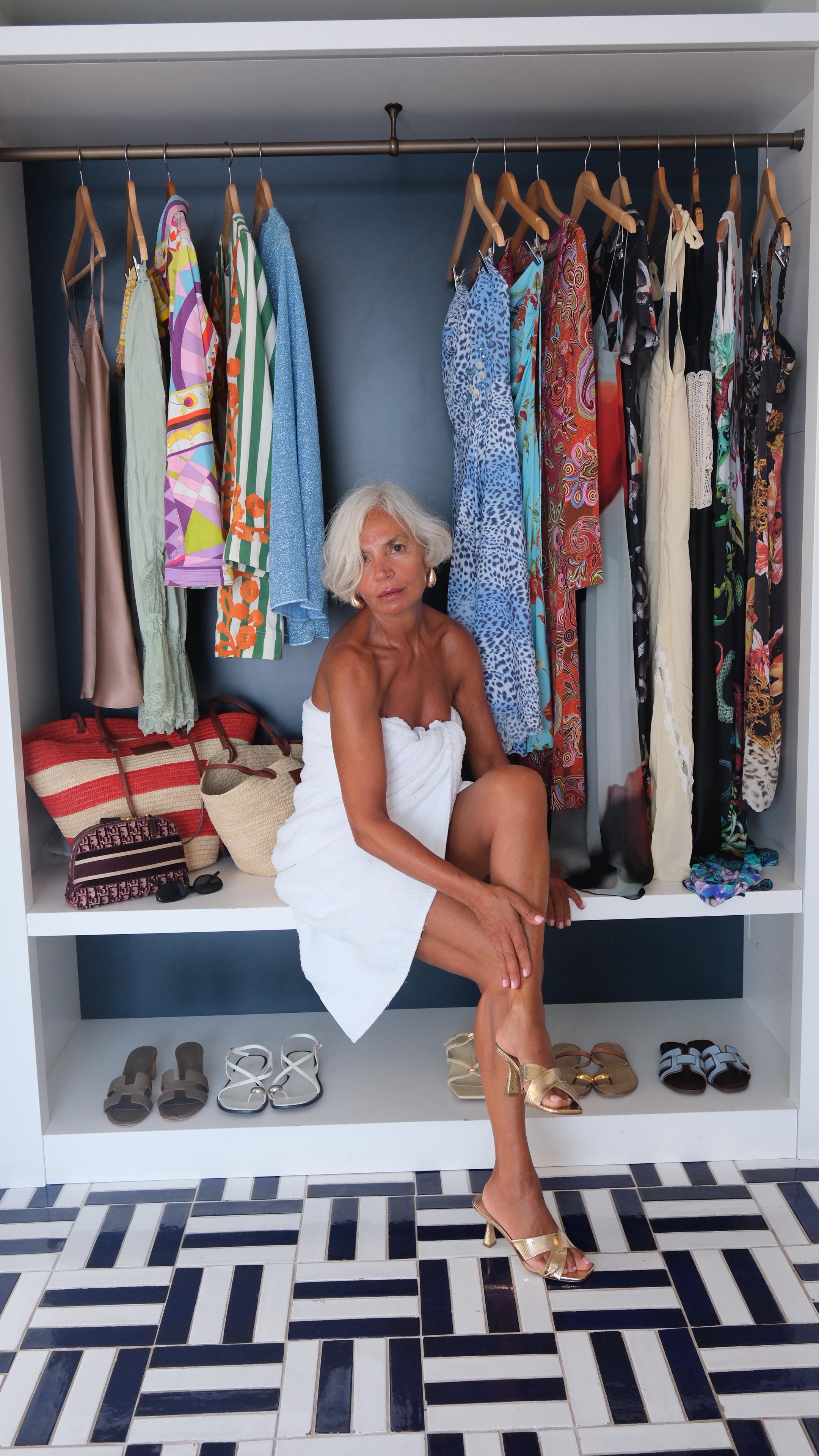 Grece Ghanem sitting in a white fluffy towel wrapped around her in front of clothing on hangers, applying body care to her limbs