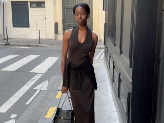 French woman wears a Khaite dress with a sweater tied around her waist, pointy toe heels and carrying a Toteme bag.